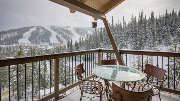 Een balkon met een glazen tafel en metalen stoelen, met uitzicht op een besneeuwd berglandschap en een skigebied in de verte.