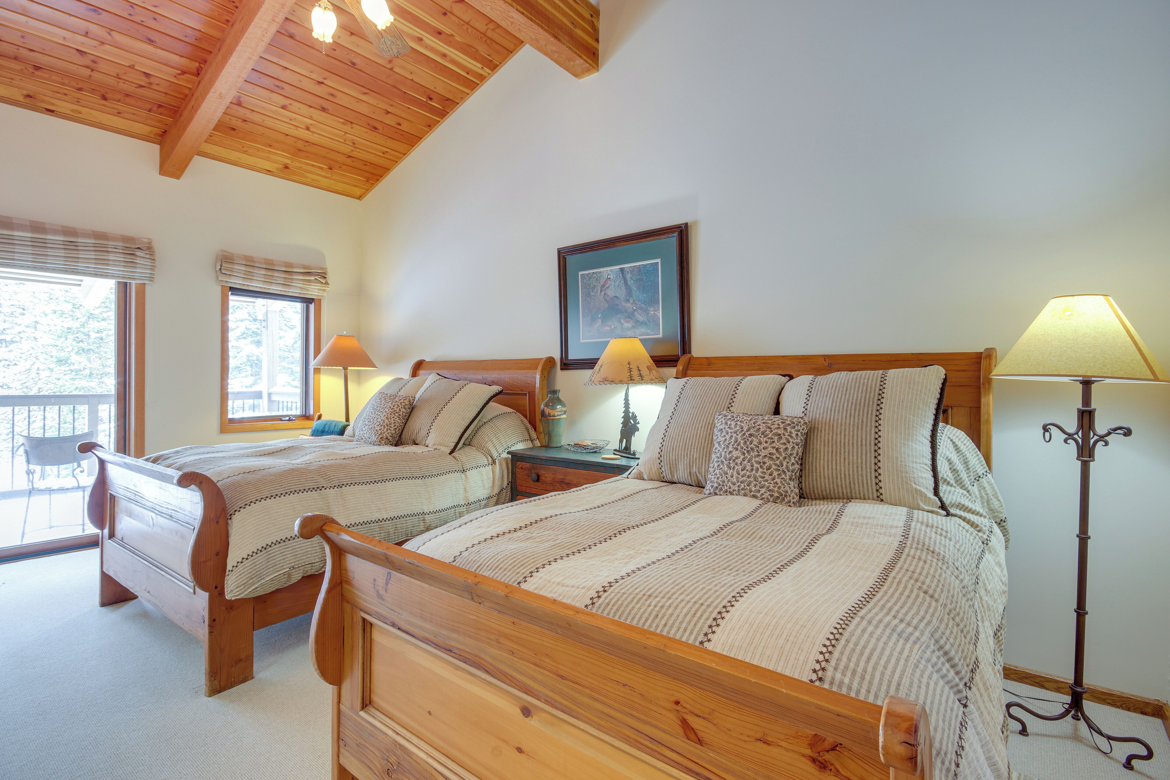 Un dormitorio con dos camas de madera, una mesita de noche con lámparas, un cuadro enmarcado en la pared y una ventana con vistas.