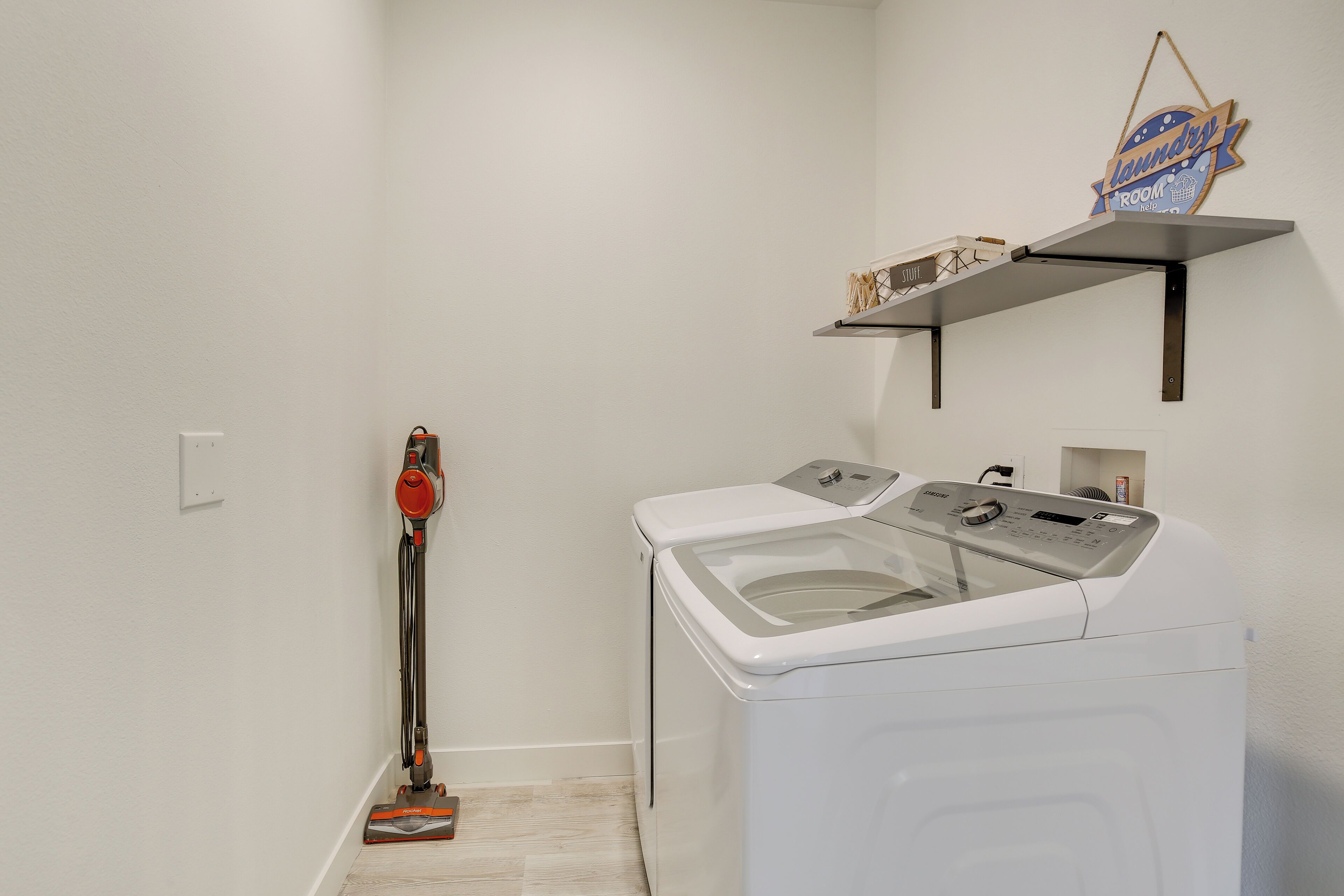 A laundry room with a washing machine and dryer, a vacuum cleaner, and a shelf with laundry supplies.