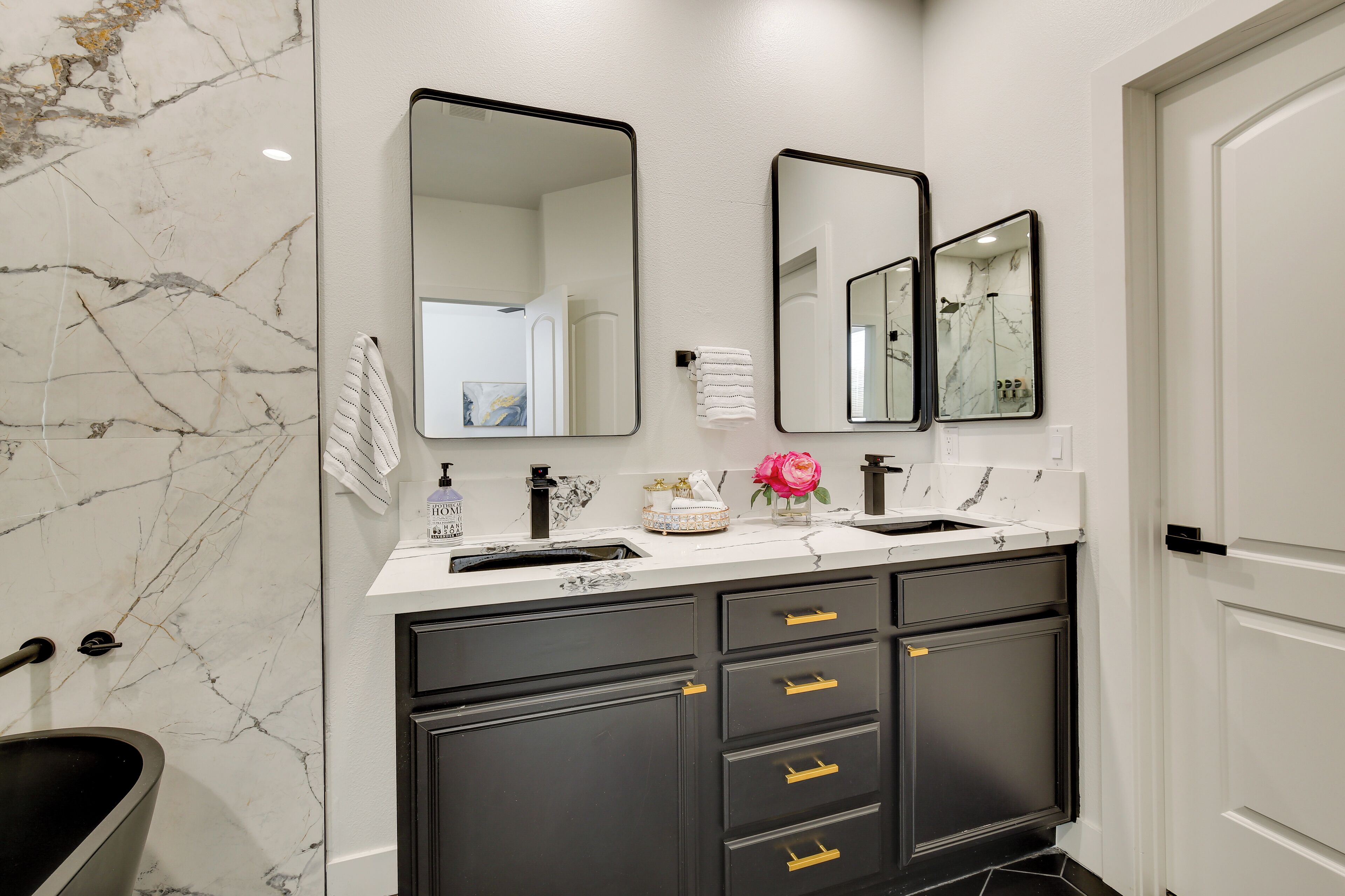 A bathroom with a marble backsplash, two sinks, a mirror, and a black vanity with gold handles.