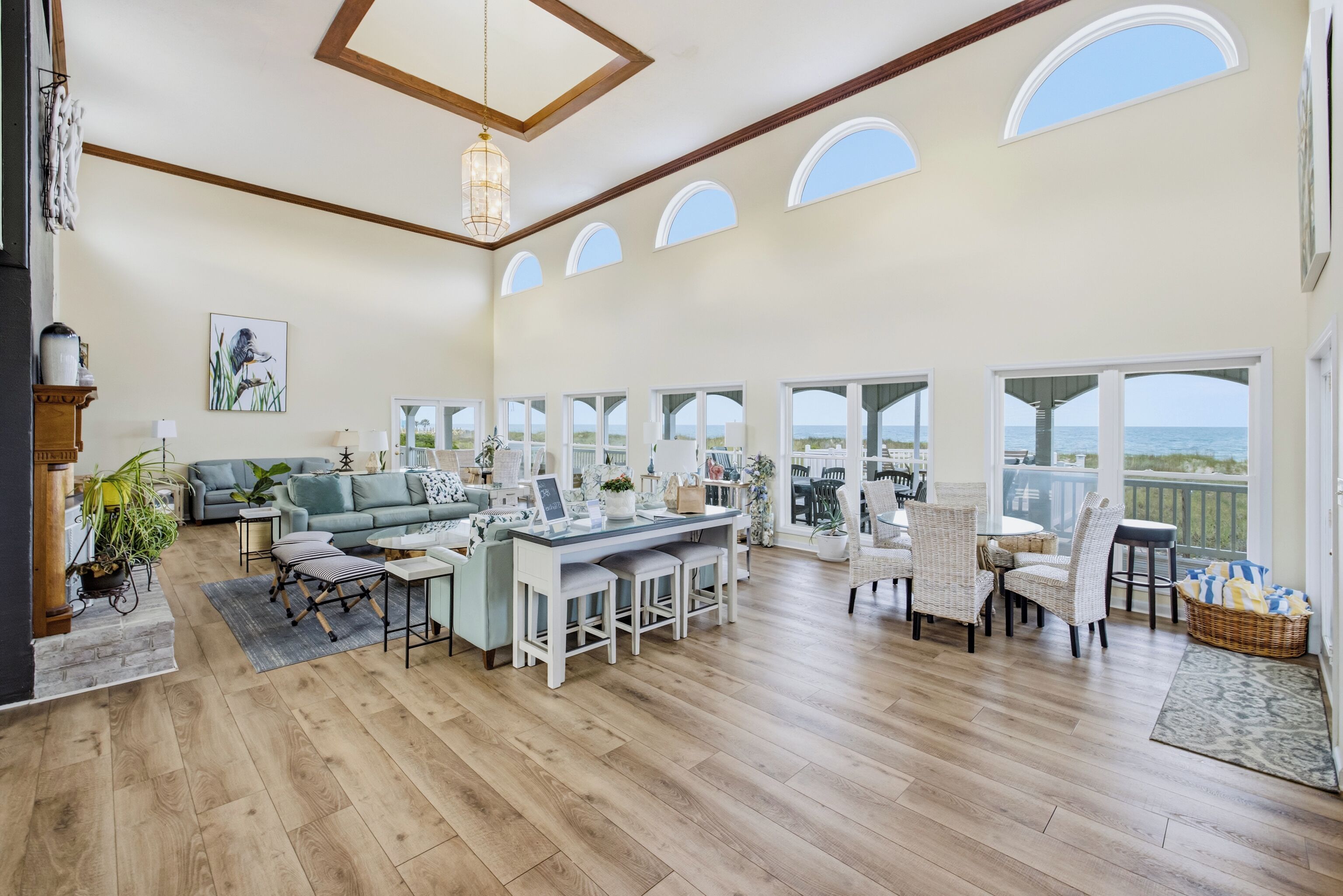 A spacious living area with a wooden floor, a sofa, a dining table with chairs, and large windows offering a view of the sea.