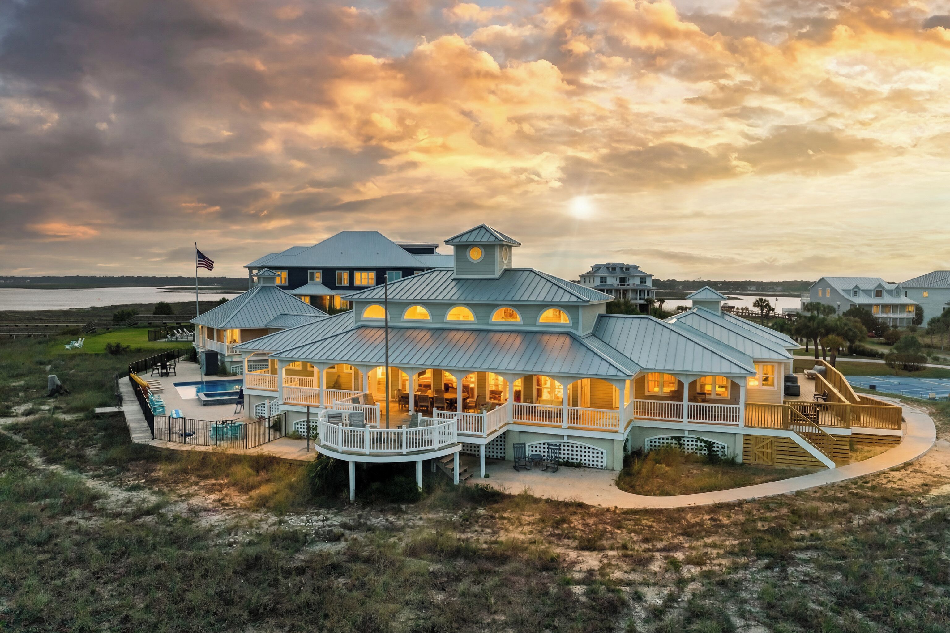A large, multi-story beachfront house with a wrap-around porch, a swimming pool, and a view of the ocean.