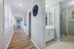 A hallway with wooden flooring, white walls, and a bathroom with a glass shower and white cabinets.