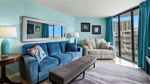Un salon avec un canapé bleu, un banc gris, un fauteuil beige et une grande fenêtre donnant sur des immeubles.