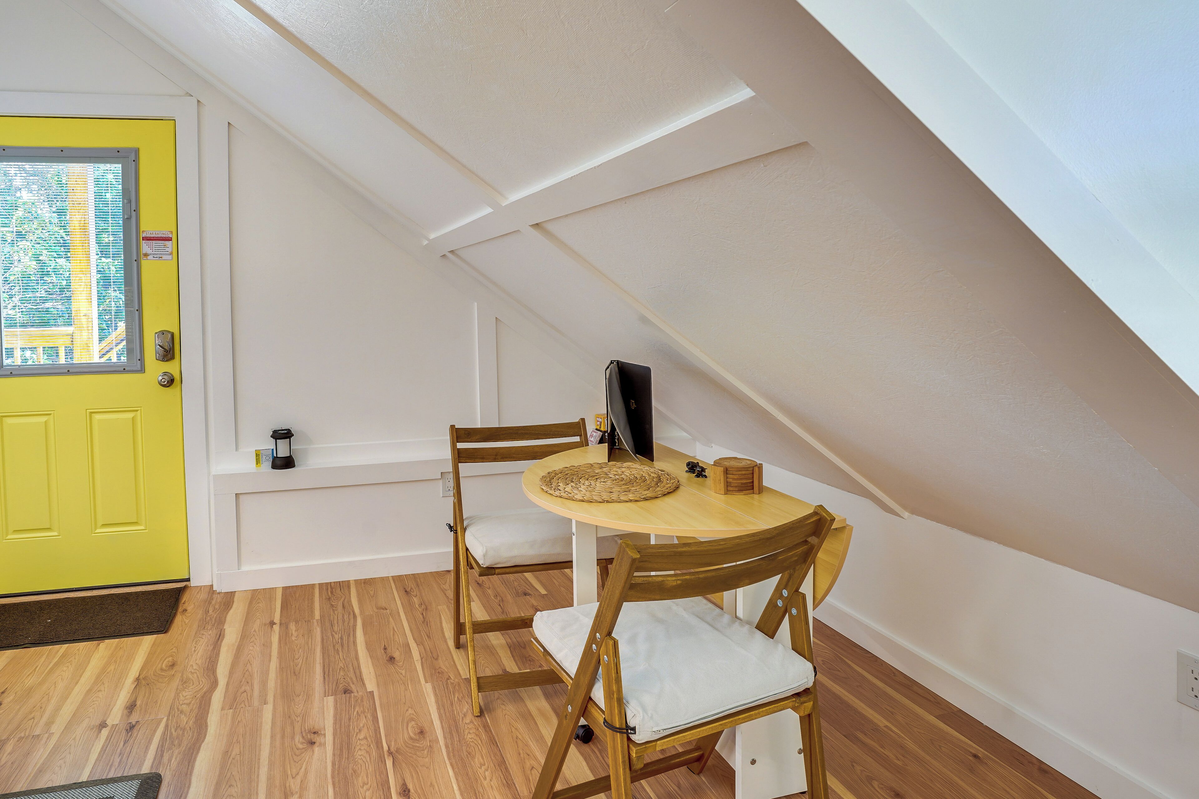 A room with a yellow door, a round wooden table with two chairs, a small shelf with a lamp, and a window with blinds.