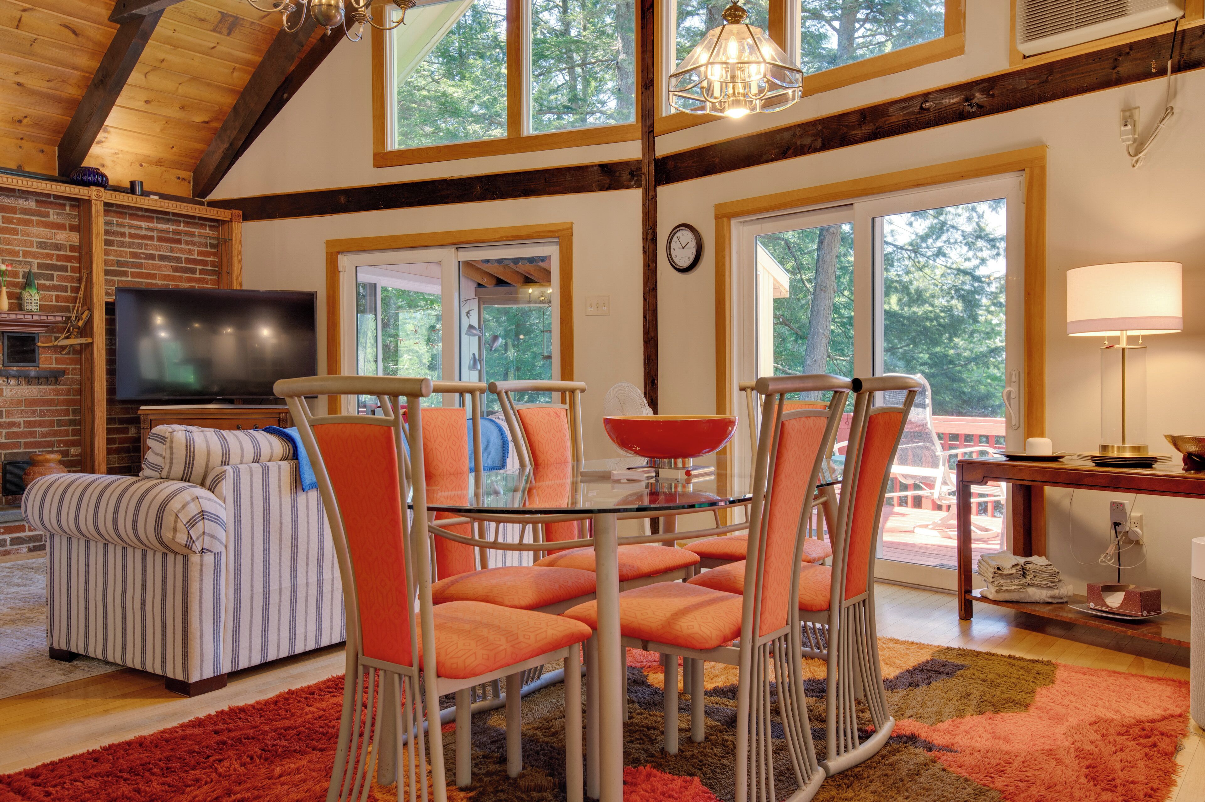 A dining area with a glass table and orange chairs, a television in the corner, and a brick fireplace.