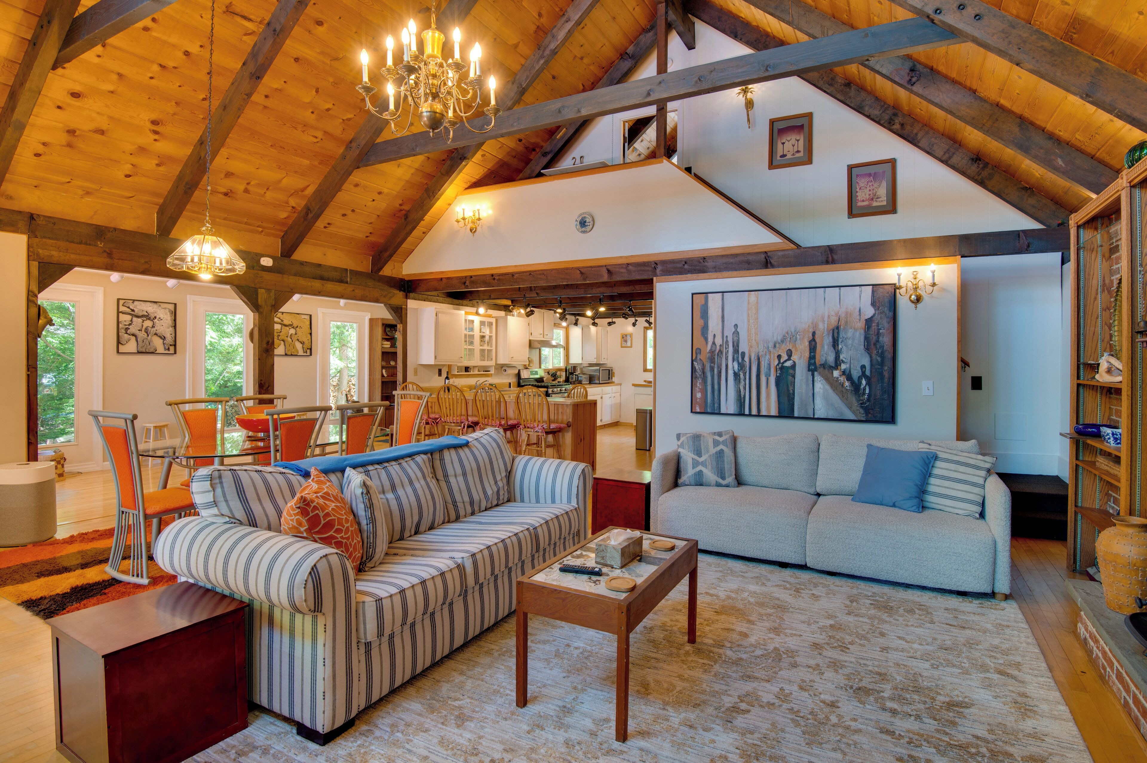 A spacious living area with a high ceiling, wooden beams, a dining area with orange chairs, and a kitchen in the background.