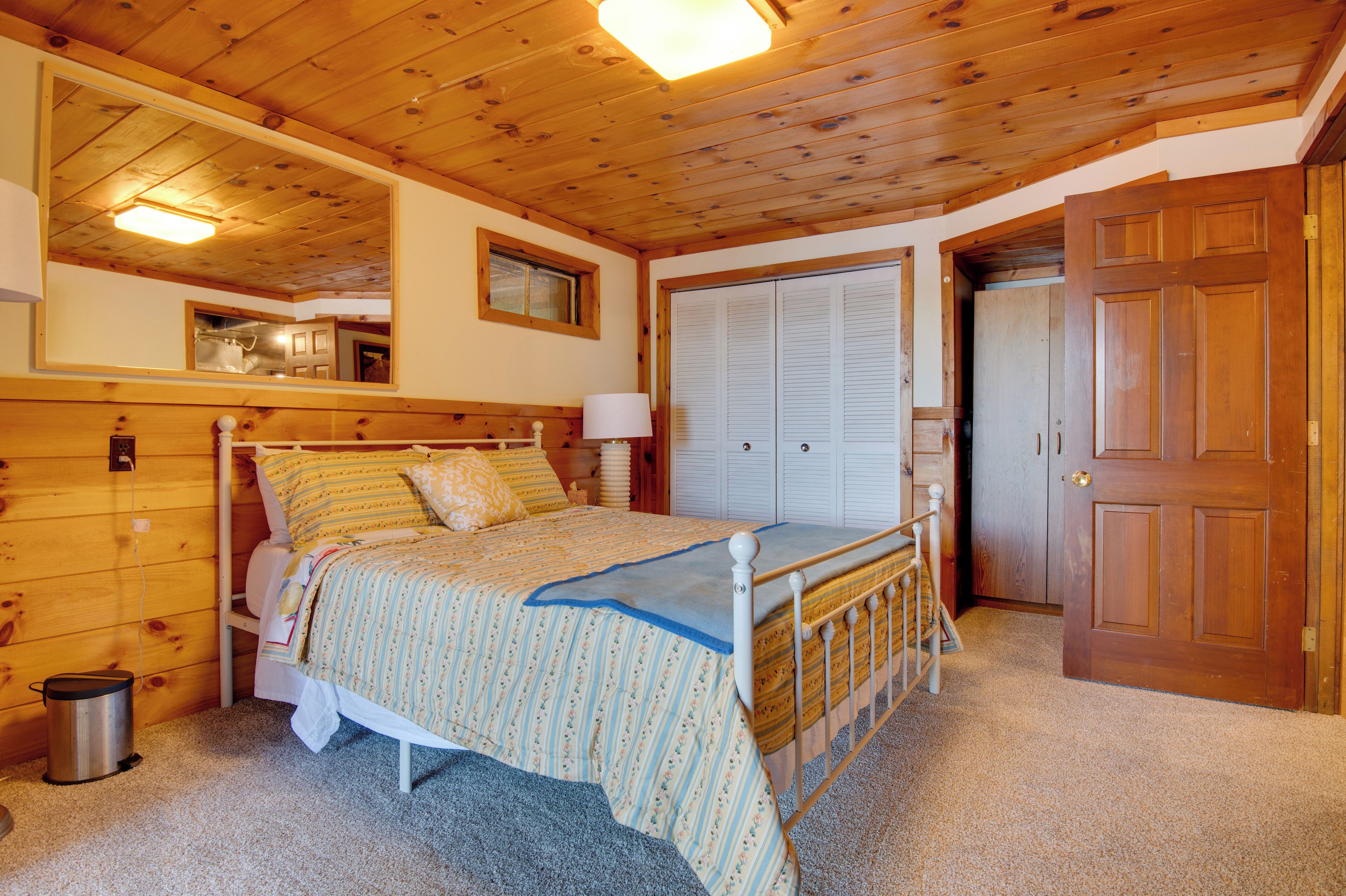 Une chambre aux murs lambrissés, avec un lit, une petite fenêtre, une porte et une poubelle.