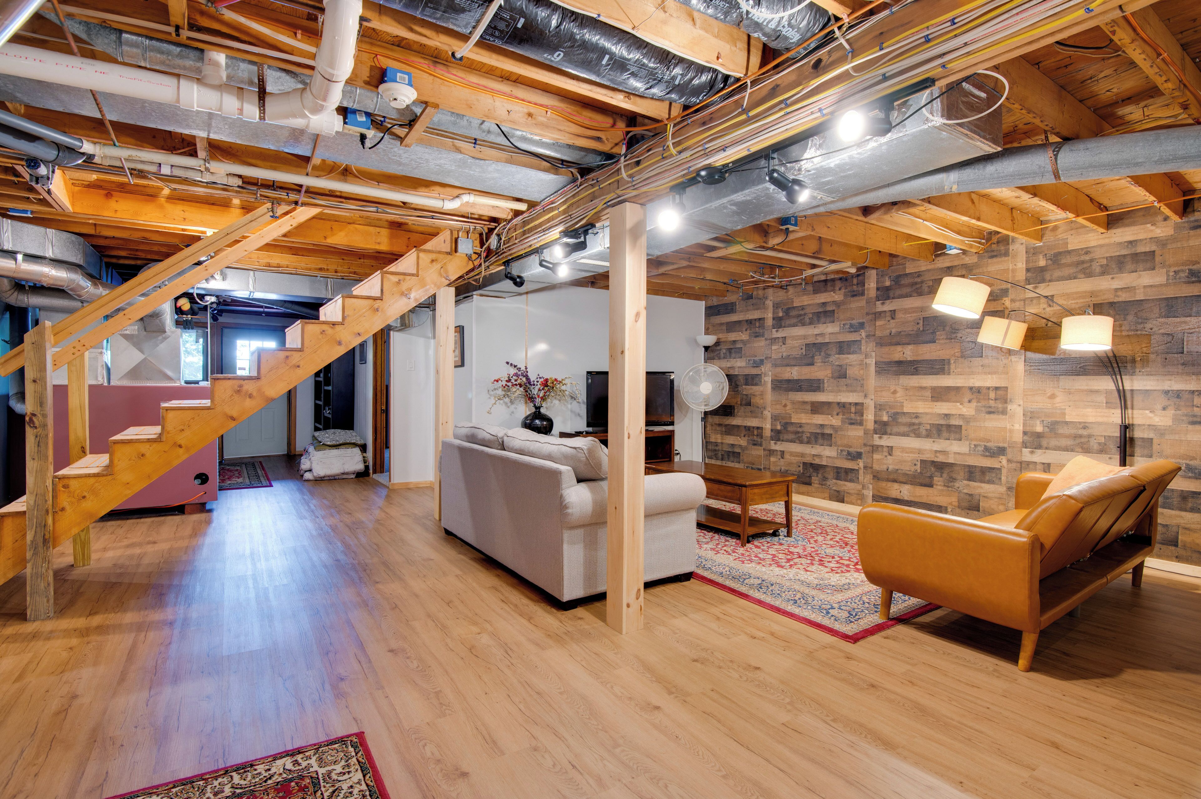 Een ruime loft met zichtbare houten balken, een trap, een witte bank, een leren fauteuil en een vloerkleed met een rood- en blauw patroon.