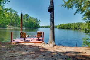 Ein Steg am See mit zwei Stühlen und einem Tisch, umgeben von Bäumen und einer ruhigen Seenlandschaft.