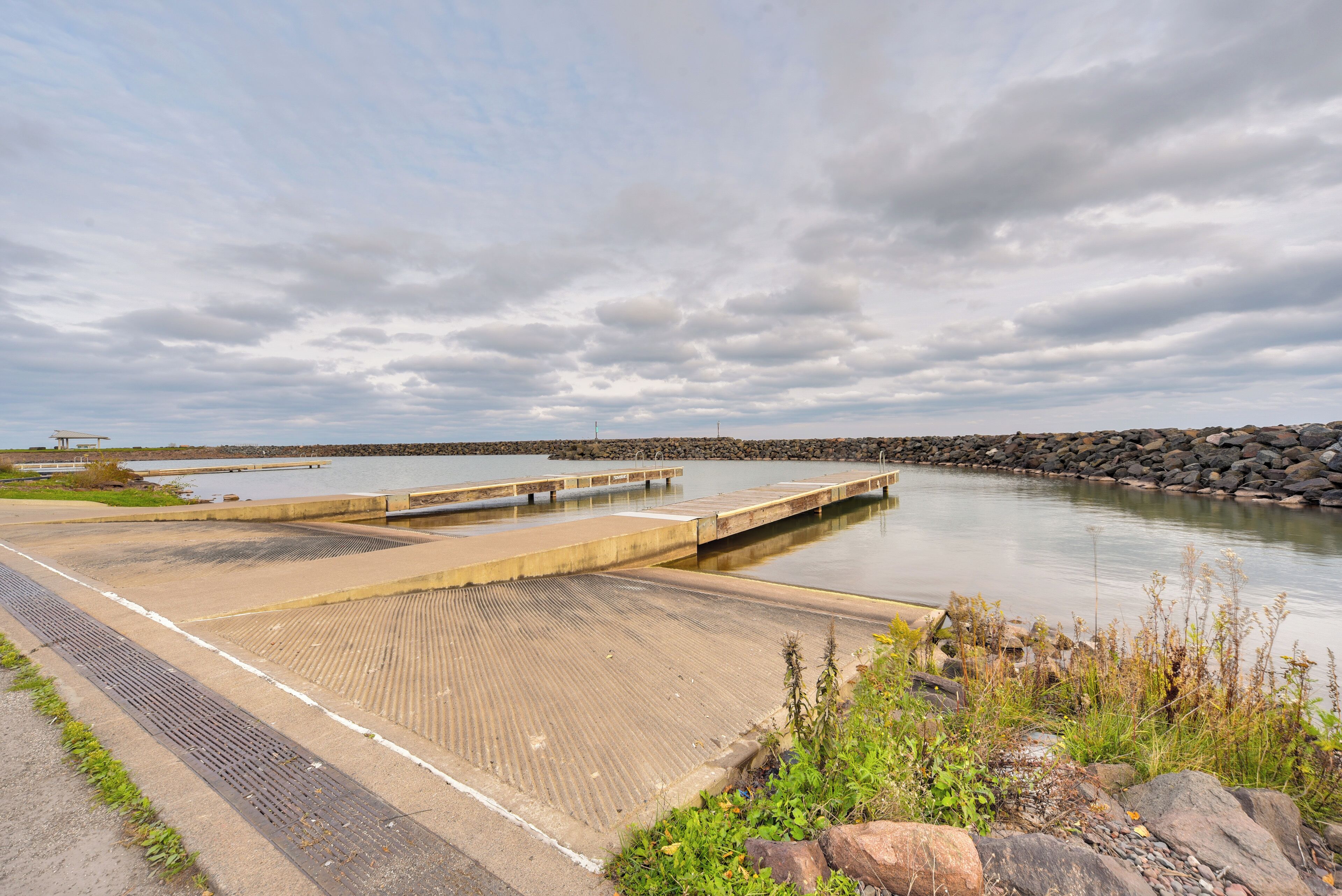 A calm waterfront area with a concrete dock extending into a body of water, surrounded by a rocky shoreline and overcast sky.