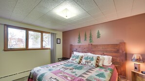 A bedroom with a large bed, a wooden headboard, a window with a view of trees, and a wall-mounted lamp. - Lake Superior Getaway - Walk to Water! (Duluth)
