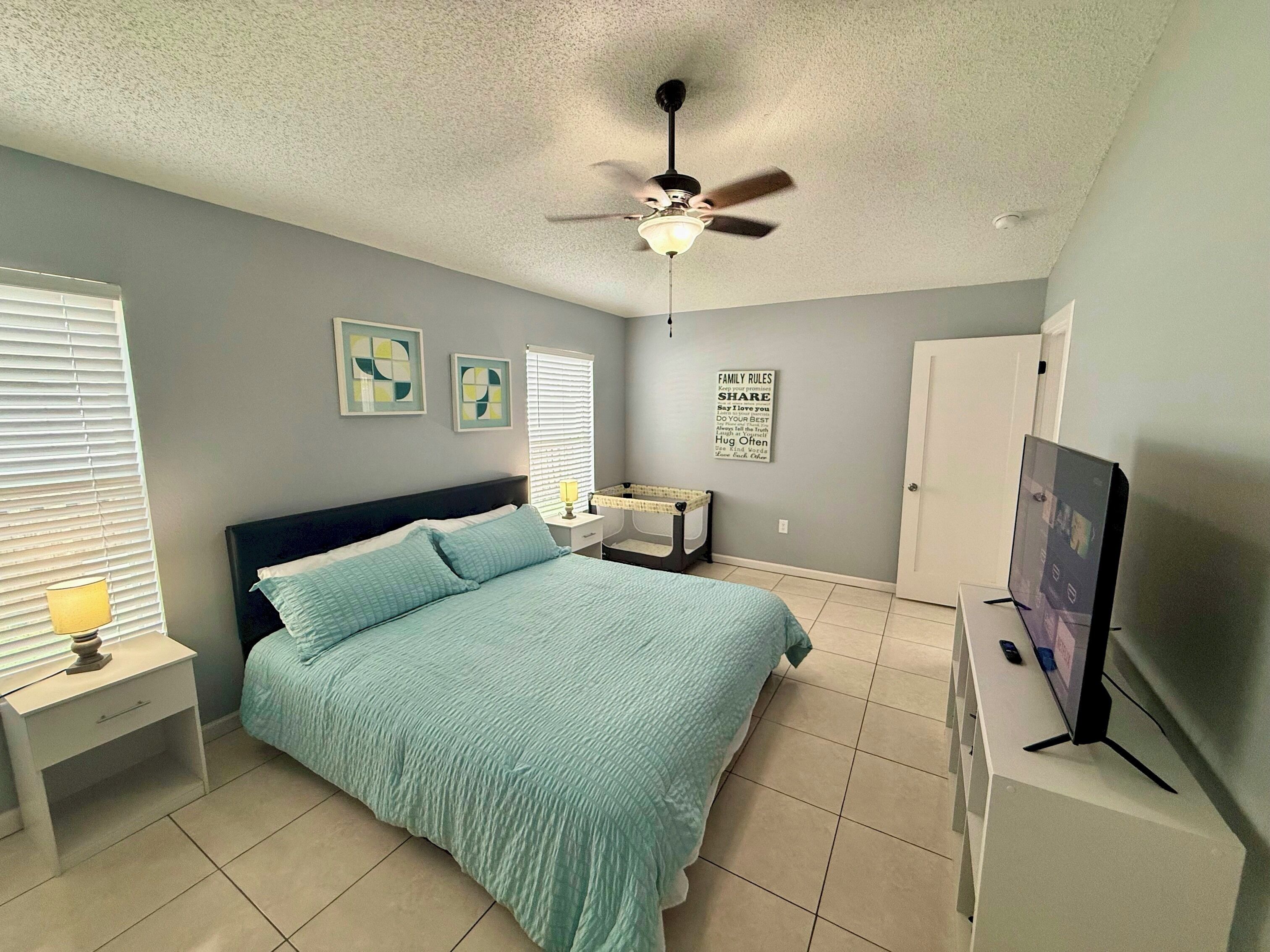 A bedroom with a bed, a ceiling fan, a TV, and a wall with a family rules poster.