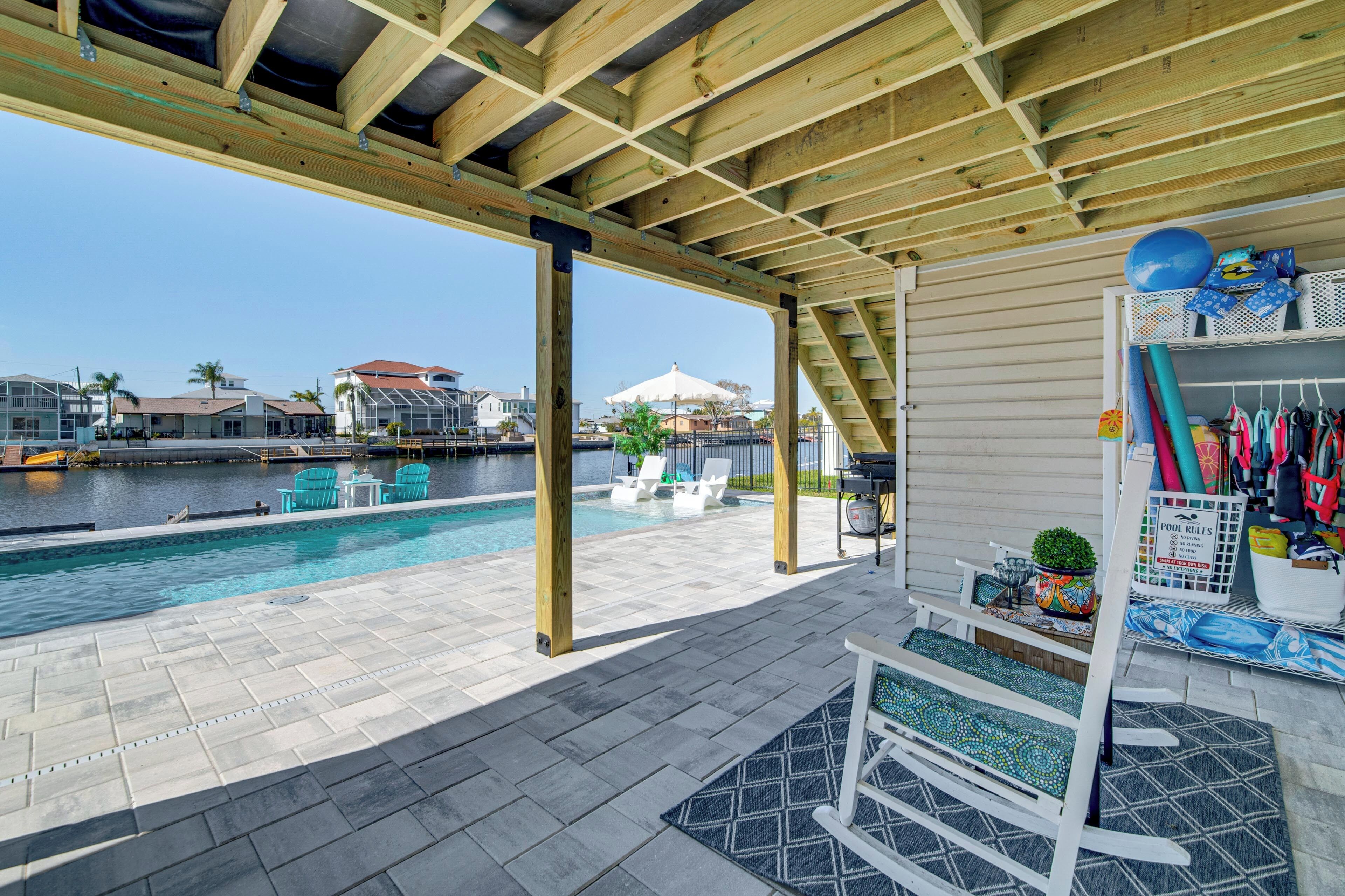 A covered pool area with a wooden ceiling, a poolside lounge with chairs, a small table, and a rack with pool accessories.