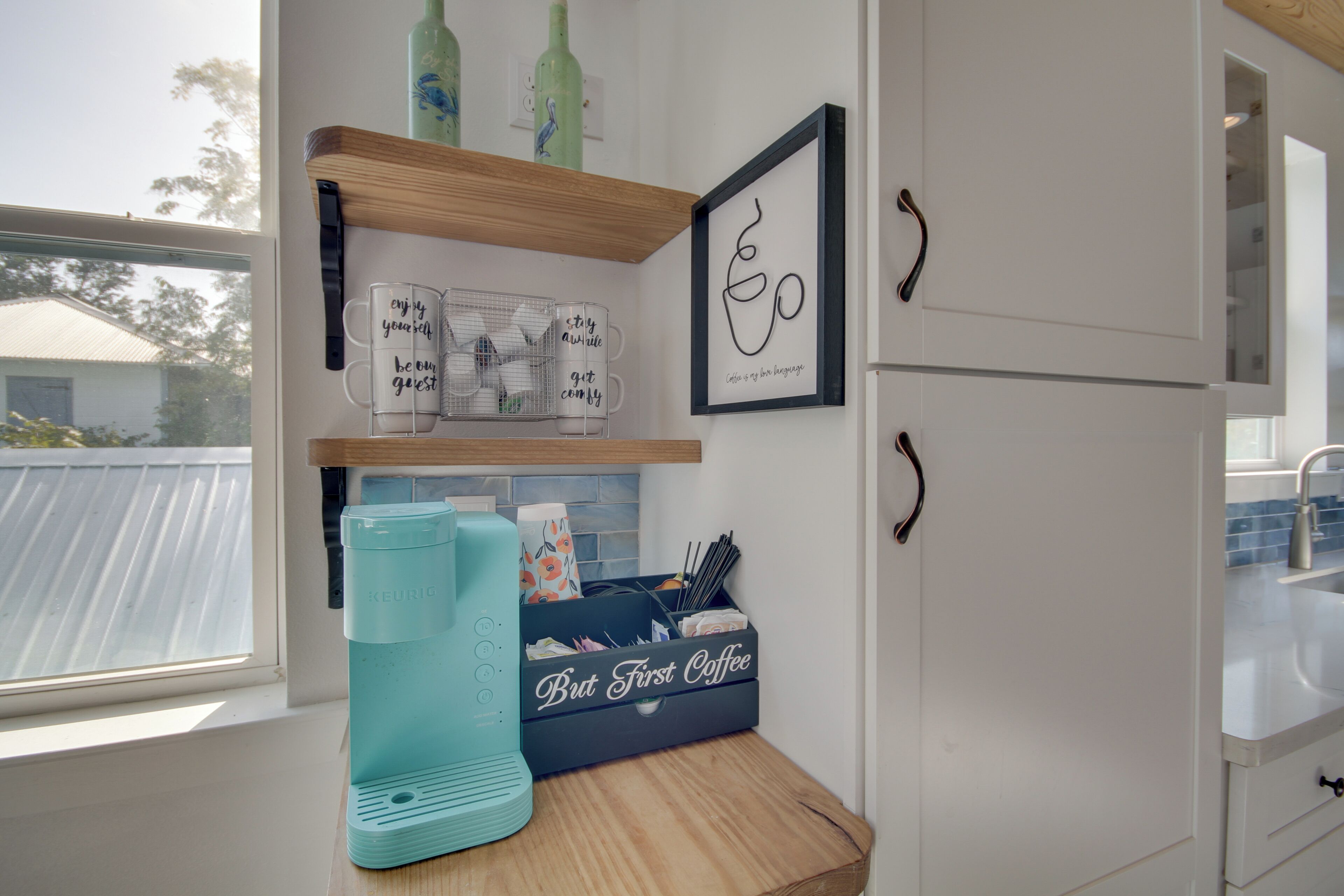 A kitchen with a window, a teal coffee maker, mugs, and a coffee-themed storage box.