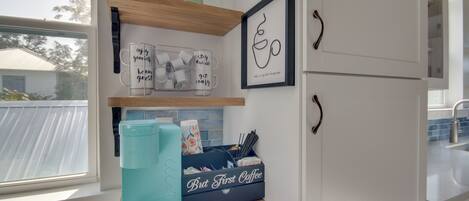 A kitchen with a window, a teal coffee maker, mugs, and a coffee-themed storage box.