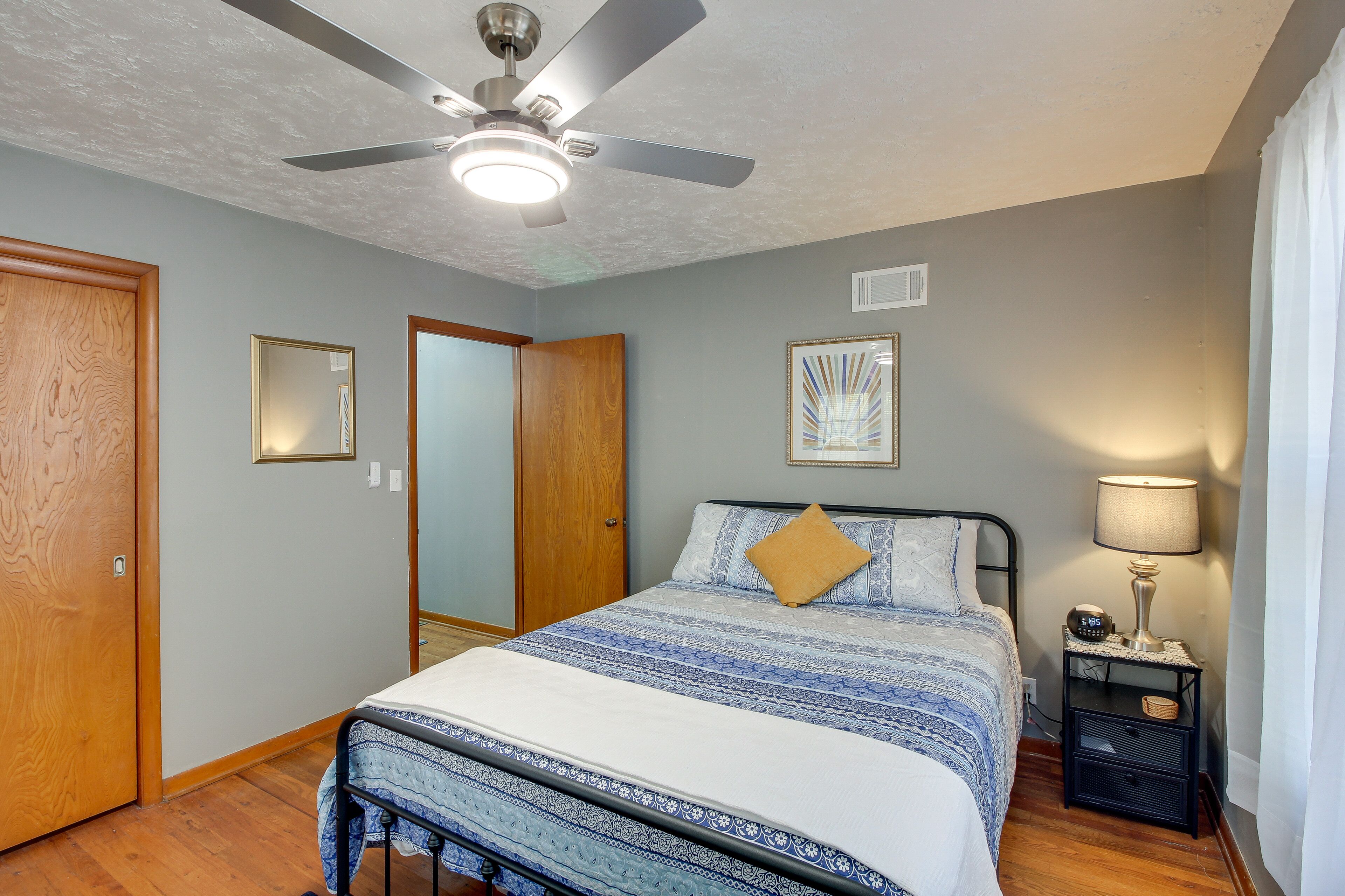 A bedroom with a bed, a ceiling fan, a nightstand with a lamp, and a mirror on the wall.