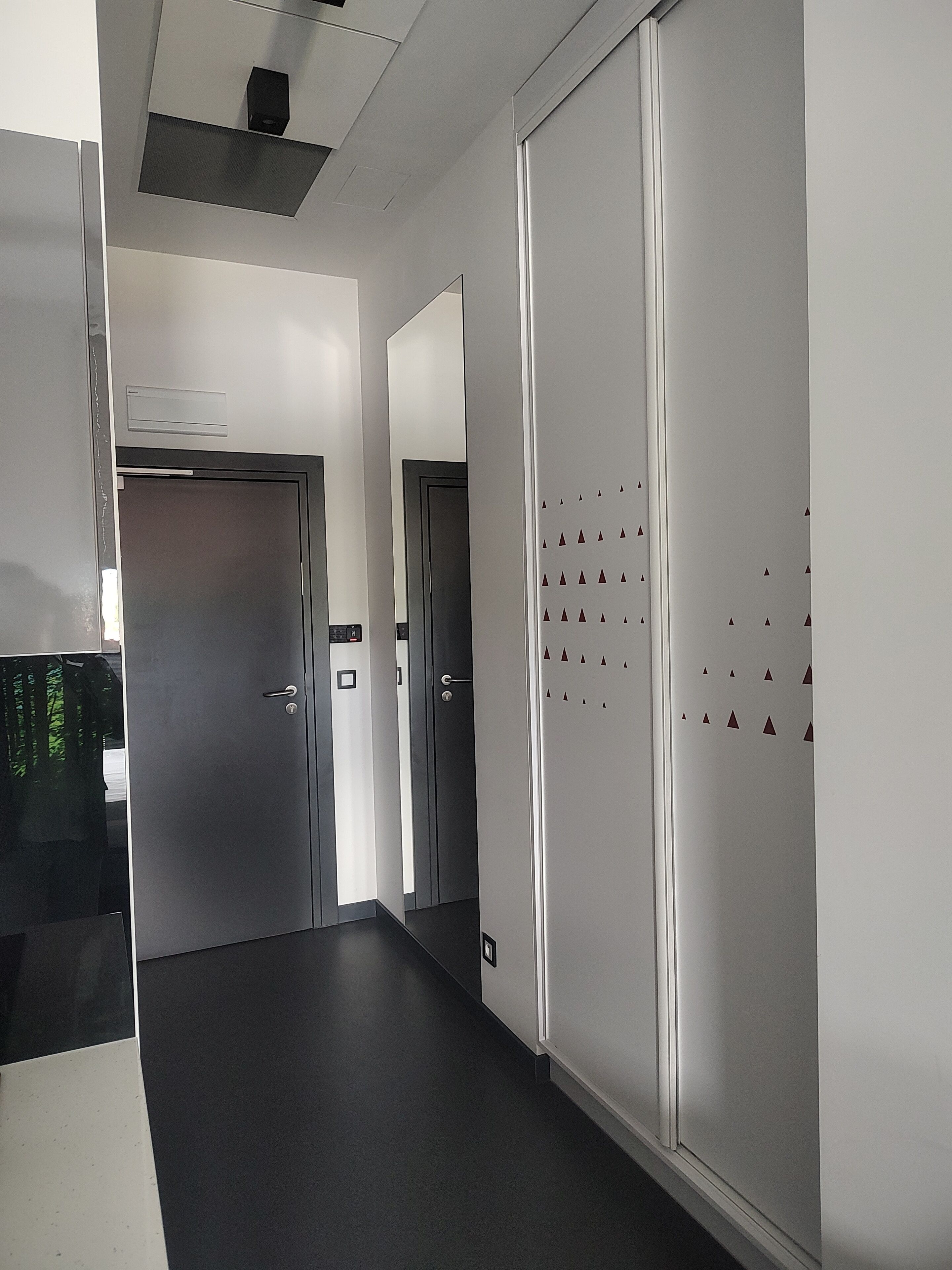 A hallway with a door at the end, a wall with a geometric pattern, and a ceiling with a vent.