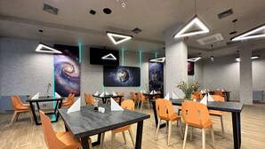 Un espace repas moderne avec des tables et des chaises orange, agrémenté de luminaires au plafond aux motifs géométriques et d’œuvres d’art murales sur le thème de l’espace.