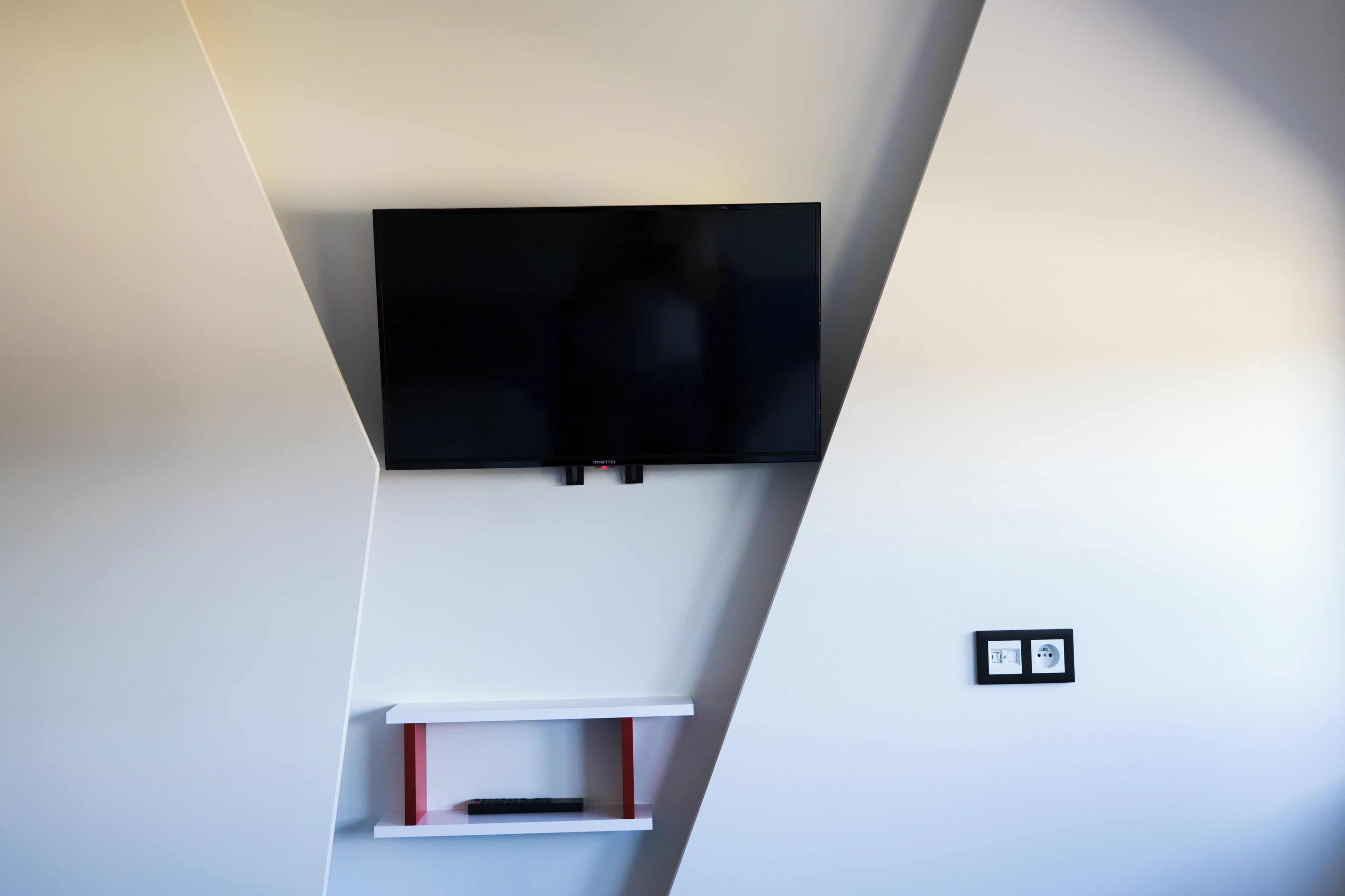 Een moderne kamer met een flatscreen televisie aan de wand, een witte plank eronder en een stopcontact aan de rechterkant.
