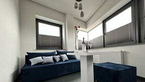 Une pièce moderne avec un canapé bleu foncé, une table blanche et un banc. Il y a deux fenêtres avec des stores, un radiateur et un vase avec des fleurs sur la table.