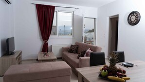A living room with a sofa, a television, a dining table with fruit, and a view through a window. - Haritos Holidays Home (Pompia)