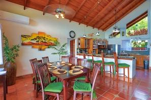 Uma sala de jantar ampla, com mesa e cadeiras de madeira, um quadro grande na parede e uma cozinha ao fundo.