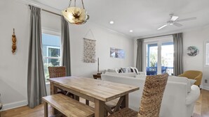 A modern living room with a wooden dining table, wicker chairs, a white sofa, and a ceiling fan. - Beach Lovers' Paradise: Beach, Pier, and Dining Nearby! (Wrightsville Beach)