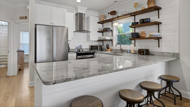 A modern kitchen with white cabinetry, a stainless steel refrigerator, a granite countertop, and wooden stools. - Beach Lovers' Paradise: Beach, Pier, and Dining Nearby! (Wrightsville Beach)