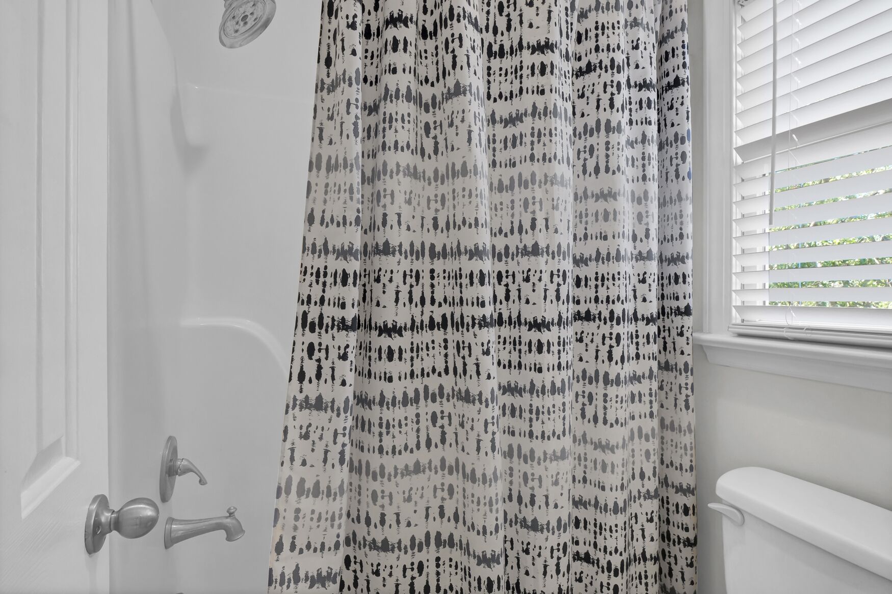 Une salle de bain avec un rideau de douche à motifs, une baignoire blanche et une fenêtre avec des stores.