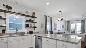 A modern kitchen with white cabinetry, a granite countertop, and a built-in dishwasher. There is a window with blue shutters, a hanging light fixture, and a view of the outdoors. - Beach Lovers' Paradise: Beach, Pier, and Dining Nearby! (Wrightsville Beach)