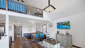 Ein geräumiges Wohnzimmer mit einer Ledersofa, einem Couchtisch und einem gemusterten Teppich. Eine Treppe führt zu einer oberen Ebene mit einem Balkon.