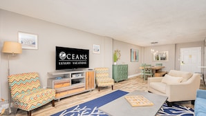 A living room with a TV displaying 'CEANS LUXURY VACATIONS', a sofa, a coffee table, and a patterned rug.