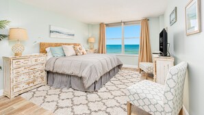A bedroom with a bed, bedside tables, a chair, a TV, and a view of the ocean.