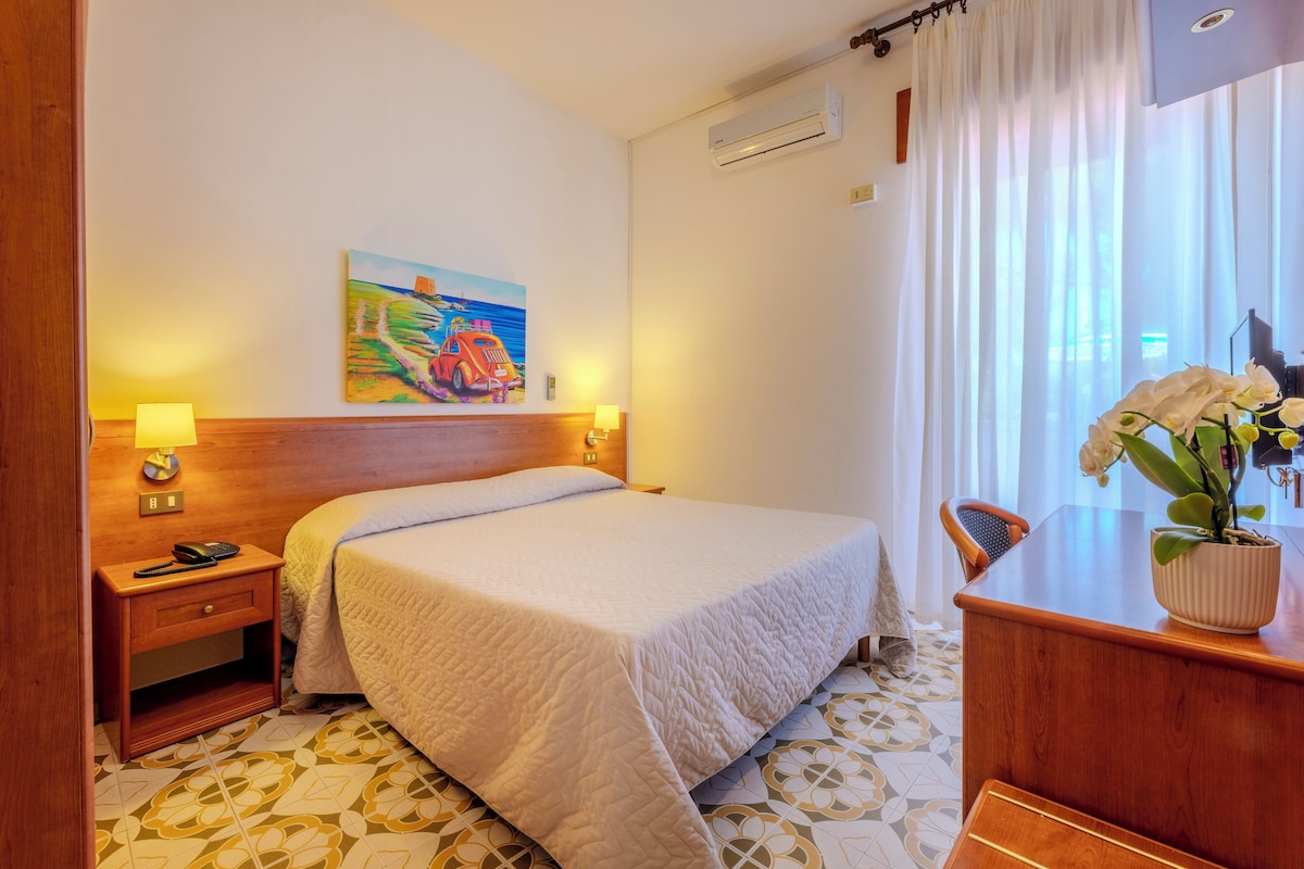 A hotel room with a bed, bedside lamps, a painting, a desk with a potted plant, and a patterned floor.