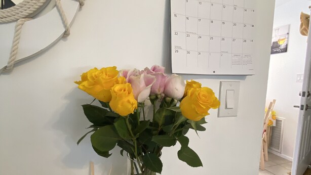 Un calendrier pour le mois de janvier, un miroir orné d’un motif de corde et un vase contenant des roses jaunes et un petit panneau portant l’inscription « aimez et vivez ».