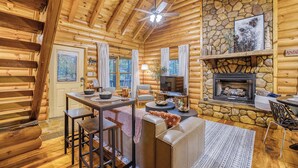 A log cabin interior with a stone fireplace, wooden walls, a dining area, and a cozy seating space. - Hot Tub · Arcade Loft · Fire Pit · Wooded Cabin (Cherry Log)