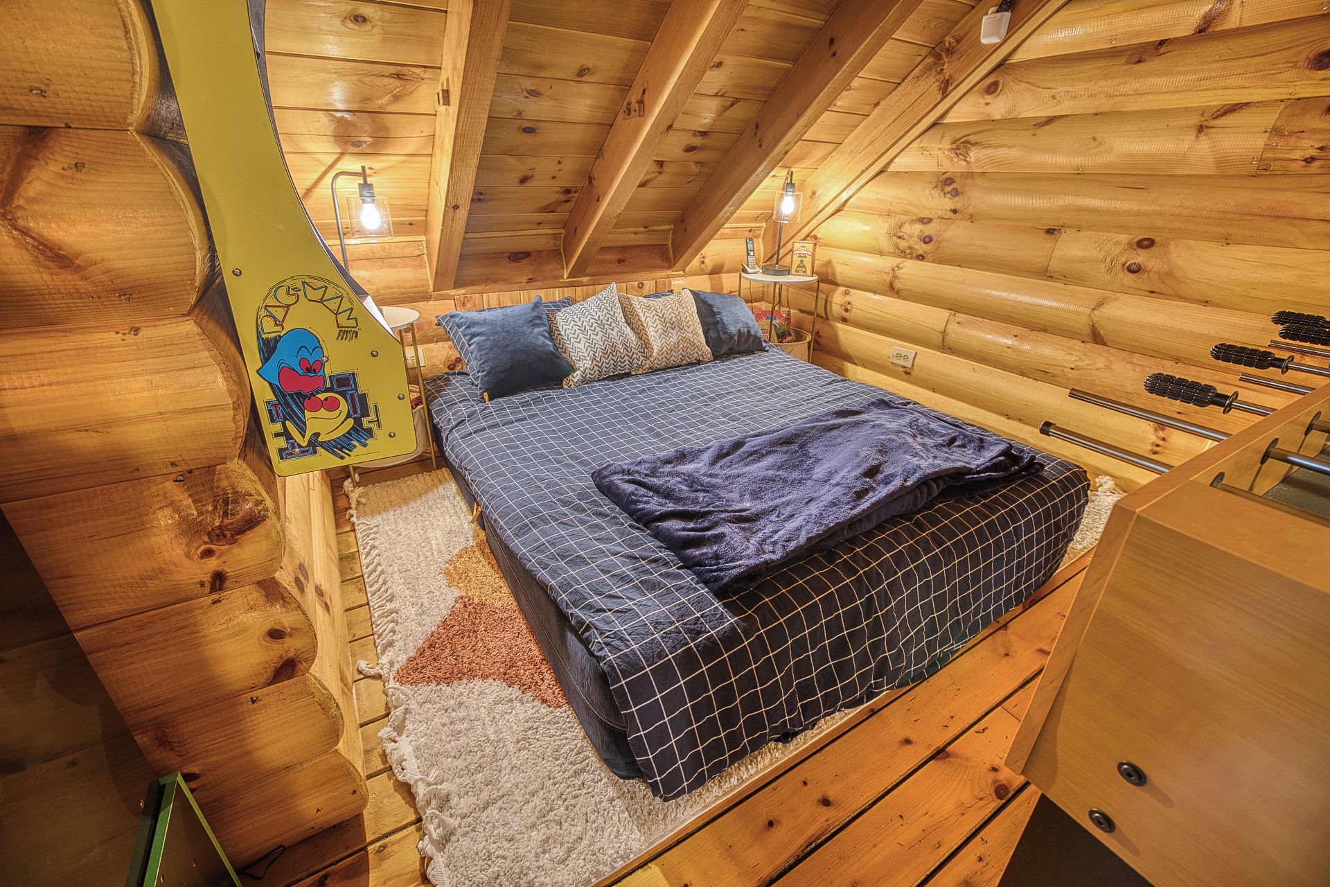 A cozy wooden cabin bedroom with a bed, pillows, a rug, and a small table with a lamp.