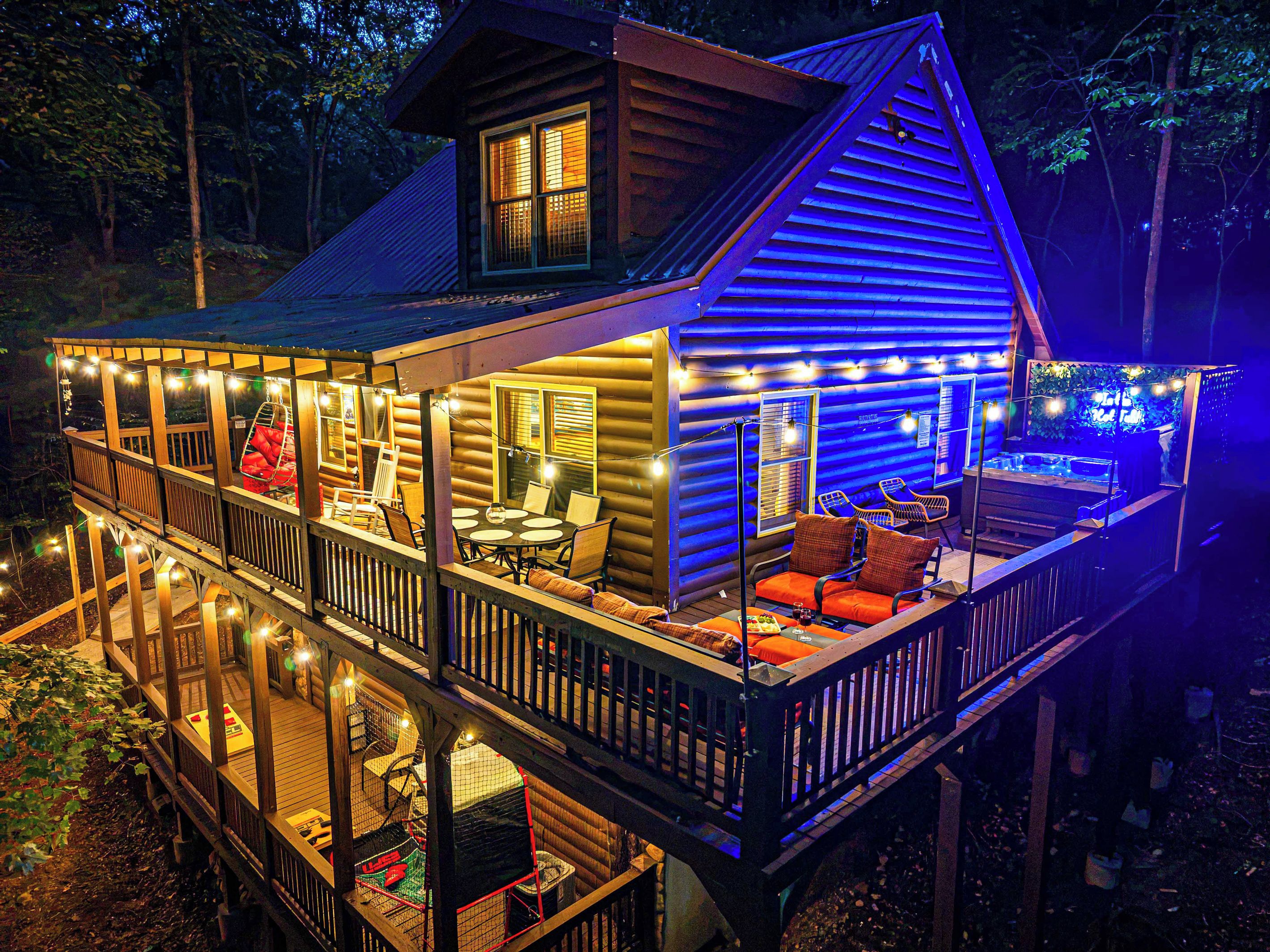 Arrive at dusk to a wraparound deck lit up with string lights. The hot tub's neon sign glows from the corner, and the whole cabin looks like it's been waiting for you.