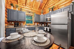A modern kitchen with grey cabinets, a stainless steel refrigerator, and a dining table set for four. - Hot Tub · Arcade Loft · Fire Pit · Wooded Cabin (Cherry Log)