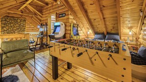 A cozy wooden cabin with a foosball table, a stone wall, and a cozy seating area. - Hot Tub · Arcade Loft · Fire Pit · Wooded Cabin (Cherry Log)