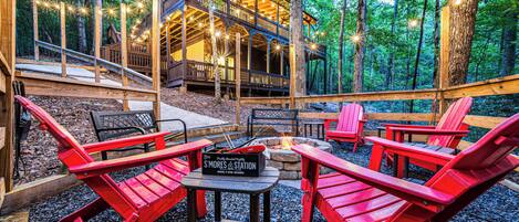 Gather the group around the fire pit and roast s'mores. The S'mores station is set out and ready, the trees close in around you, and the cabin sits up the hill.