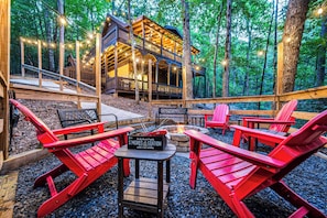 Gather the group around the fire pit and roast s'mores. The S'mores station is set out and ready, the trees close in around you, and the cabin sits up the hill. - Hot Tub · Arcade Loft · Fire Pit · Wooded Cabin (Cherry Log)