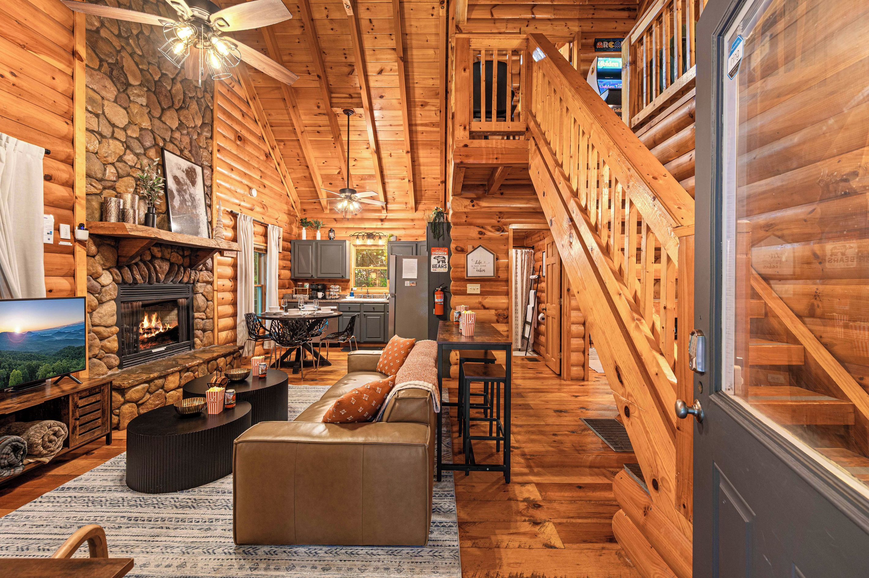 A log cabin interior with a stone fireplace, a television, a sofa, and a staircase leading to a loft.
