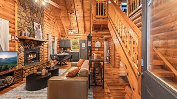 A log cabin interior with a stone fireplace, a television, a sofa, and a staircase leading to a loft.