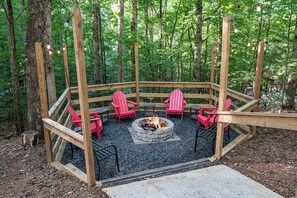 A circular wooden deck with a fire pit in the center, surrounded by red chairs, set in a forested area. - Hot Tub · Arcade Loft · Fire Pit · Wooded Cabin (Cherry Log)