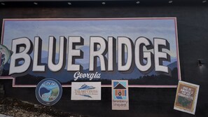 A mural with the words "BLUE RIDGE Georgia" and logos for the City of Blue Ridge, The Art Center Blue Ridge Mountains Arts Association, and Sereberry Vineyards. - Hot Tub · Arcade Loft · Fire Pit · Wooded Cabin (Cherry Log)