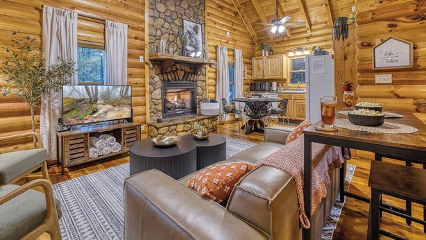 A cozy log cabin interior with a stone fireplace, wooden walls, a dining area, and a kitchen. - Hot Tub · Arcade Loft · Fire Pit · Wooded Cabin (Cherry Log)