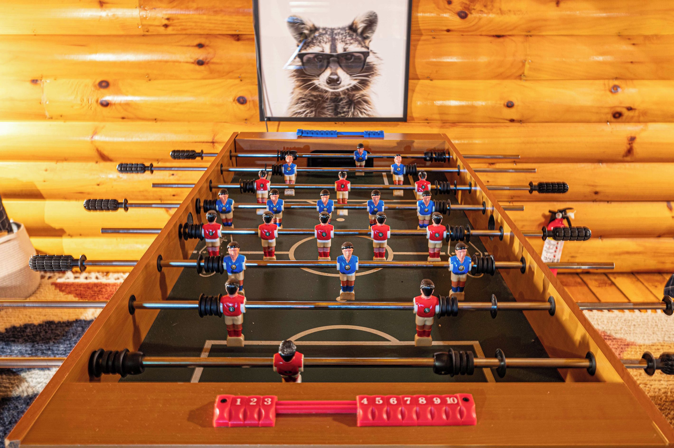 Une table de baby-foot dans une pièce aux murs en bois, avec un tableau encadré représentant un raton laveur.
