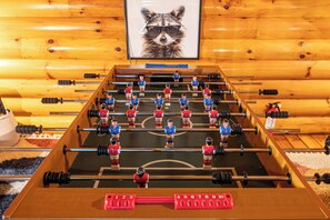A foosball table in a room with wooden walls and a framed picture of a raccoon. - Hot Tub · Arcade Loft · Fire Pit · Wooded Cabin (Cherry Log)